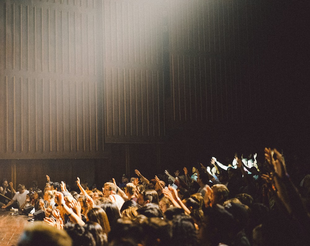 photo of a crowd in a dark room with a beam of light shining onto a stage. some people in the crowd are raising their hands.